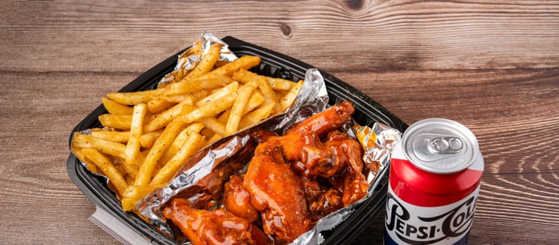 Takeout container with golden french fries, saucy chicken wings, and a Pepsi can on a wooden table.